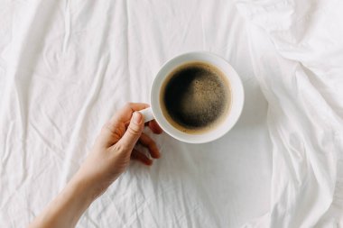 Beyaz çarşafta bir fincan kahve tutuyor. Yatak konseptinde kahvaltı.