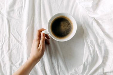 Beyaz çarşafta bir fincan sade kahve tutuyor. Yatak konseptinde kahvaltı.