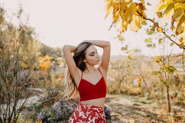 Uzun sarı dalgalı saçlı genç bir kadın, kırmızı bir bluz ve etek giyiyor, parkta..