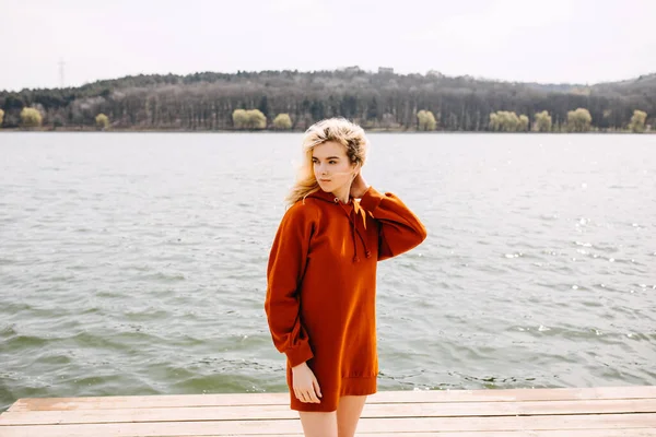 年轻的金发女郎穿着长长的红色帽衫 站在湖边迎风飘扬 — 图库照片