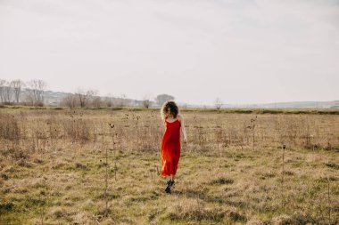 Kıvırcık saçlı genç bir kadın, açık havada kırmızı bir yaz elbisesi giyiyor, tarlada yürüyor..