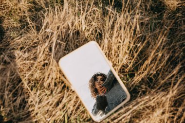 Portrait of a young woman with curly hair, reflected in a mirror placed on grass in a field. clipart