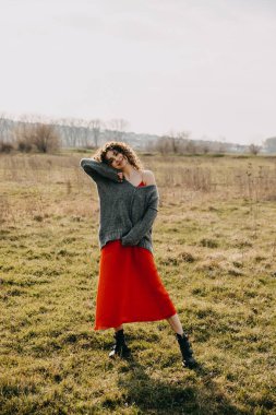 Kıvırcık saçlı, büyük boy bir kazak ve açık havada kırmızı bir elbise giyen mutlu bir genç kadın..