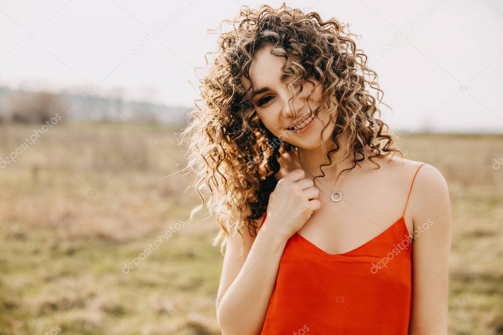 Young woman with curly hair, smiling, outdoors at sunset, looking at camera.