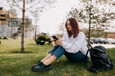 Parkta çimlerde oturan genç kız öğrenci, akıllı telefonuna bakıyor..