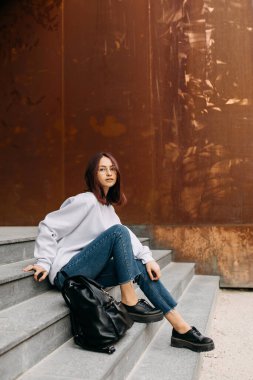 Sırt çantalı genç bir kadın beton merdivenlerde oturuyor, dışarıda..