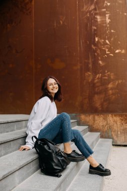 Gözlüklü kız öğrenci, gülümsüyor, üniversite binasının önündeki beton merdivenlerde oturuyor..