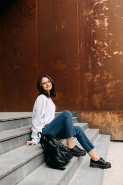 Gözlüklü, sırt çantalı, üniversite binasının önündeki beton merdivenlerde oturan bir kız öğrenci..