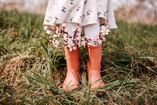 Closeup of a little girl standing in a garden with cherry tree flowers in rubber boots.