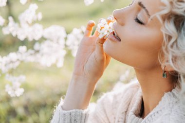 Dudaklarının yanında kiraz ağacı çiçekleri tutan kıvırcık saçlı sarışın bir kadının yakın plan portresi. Güzellik konsepti.