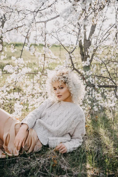 若いブロンドの髪の女性は セーターを着て 満開の桜の木の下に横たわっていた — ストック写真