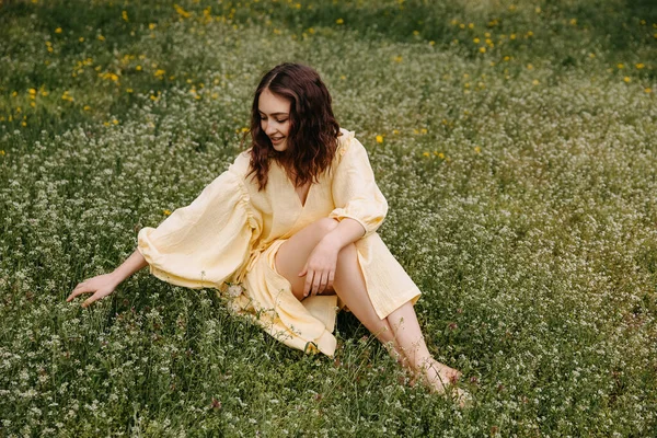 若い裸足の女性は黄色の夏のドレスを着て 緑の芝生のフィールドに座って 笑みを浮かべて — ストック写真