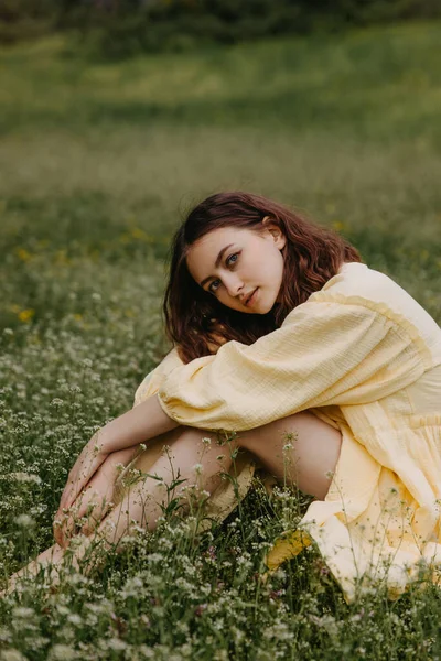 若い女性は黄色の夏のドレスを着て 緑の芝生のフィールドに座って カメラを見て — ストック写真
