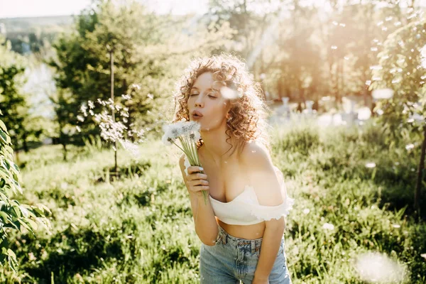 Young woman with curly hair, blowing dandelions in a park on summer day.