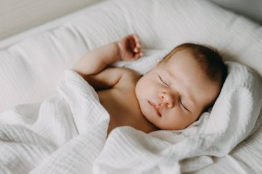 Yeni doğmuş bebek, beyaz battaniyeyle kaplı bir bebek yuvasında uyuyor..