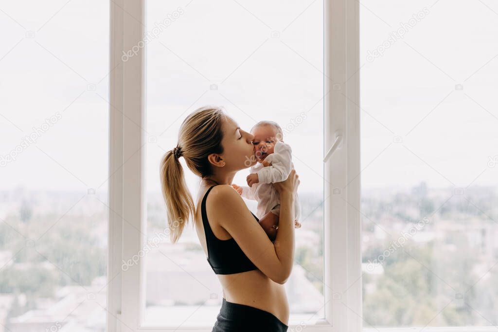 Young mother holding newborn baby in her arms at home, by the window, kissing her on cheek.