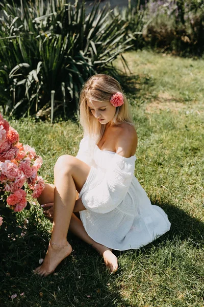 在一个阳光灿烂的夏天 年轻的金发女子坐在绿草上 旁边是一丛丛粉红色的玫瑰 穿着一件白色的连衣裙 — 图库照片
