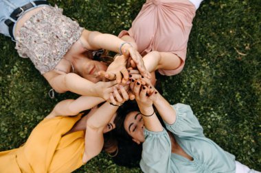 Group of four young women lying on green grass holding hands up in the air.