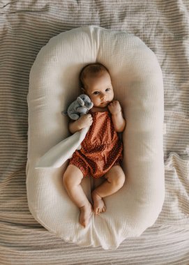 Üç aylık bebek bir kız bebek yuvasında yatıyor, pelüş oyuncak bir file sarılıyor..