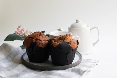 Sıcak, davetkar bir sahne. Siyah kağıda sarılı altın sarısı bir kek beyaz bir çaydanlık ve çay fincanının yanında.