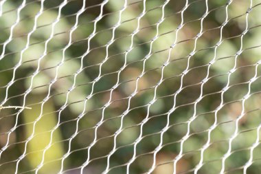 Birbirine dolanmış metal kabloların yinelenen deseni, çerçeveye uzanan ışığı yakalayarak hassas bir ağ oluşturur. Bulanık yeşil arka plan derinlik ve kontrastı ekler, sıradan bir yapısal elementi sakin ve soyut bir bileşime dönüştürür.