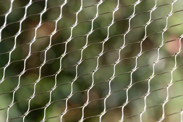 Birbirine dolanmış metal kabloların yinelenen deseni, çerçeveye uzanan ışığı yakalayarak hassas bir ağ oluşturur. Bulanık yeşil arka plan derinlik ve kontrastı ekler, sıradan bir yapısal elementi sakin ve soyut bir bileşime dönüştürür.