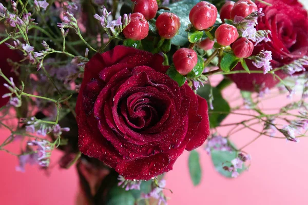 Taze çiğ damlalarıyla süslenmiş koyu kırmızı güller, parlak kırmızı hiperikum meyveleri ve narin pembe dolgu çiçekleriyle çevrili yakın plan bir çiçek sergisi..