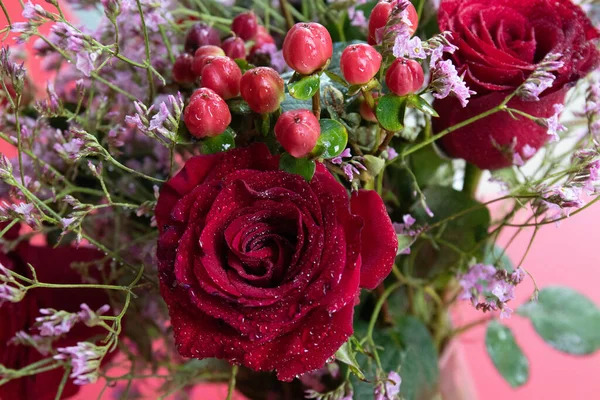 Taze çiğ damlalarıyla süslenmiş koyu kırmızı güller, parlak kırmızı hiperikum meyveleri ve narin pembe dolgu çiçekleriyle çevrili yakın plan bir çiçek sergisi..
