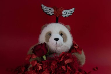 Şık bir Panda koyu kırmızı arka planda, parlak kanatlı bir kafa bandıyla süslenmiş ve koyu kırmızı gül yapraklarıyla çevrili..