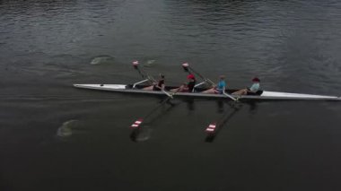 Kürek çeken bir ekip sakin bir su kütlesi üzerinde pratik yapıyor. Tek sıra halinde oturan dört kürekçi teknelerini ileri itmek için birlikte çalışıyorlar. Manzara yukarıdan, kayıkların ilerlemesini ve etrafındaki suyu kaydediyor..
