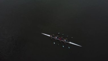 Bir kürek takımı sakin bir gölde süzülüyor, kürekleri uyum içinde hareket ediyor. Tekne durgun suyu keser ve yoluna küçük bir uyanış bırakır. Gökyüzü bulutlu, sakin atmosfere katkı sağlıyor..