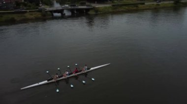 Bir kürek takımı bir nehrin sakin sularında süzülür, kürekleriyle birlikte yüzeyi keserler. Çevreleyen ağaçlar ve yemyeşil alanlar bu sakin sahne için resim gibi bir zemin oluşturuyor..