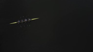 Parlak sarı bir kayıktaki bir kürek ekibi alacakaranlıkta sakin bir gölde süzülür, senkronize vuruşları karanlık sularda dalgalanmalar bırakır..