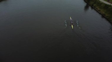 Şafak sökerken, mürettebat zarif bir şekilde Gent 'in durgun sularında yüzüyor, senkronize vuruşları huzurlu nehirde dalgalanmalar yaratıyor. Sabahın erken saatlerinin güzelliği manzarayı sarar..