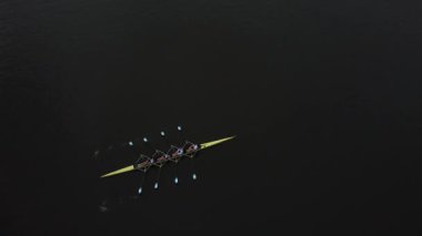 Gün batımında sakin bir gölün yüzeyinde süzülen bir kürek ekibi kürek çekerken uyum içinde kürek çekerken hafif dalgalar oluşturuyor..