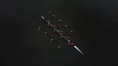 Sakin bir gölün yüzeyinde süzülen kürek takımının hava görüntüsü. Güneş suyun üzerinde parlıyor, tekneyi ve kürekçileri aydınlatıyor. Sekiz kürekçinin hepsi senkronize, kürekleri ritmik hareket ediyor..