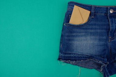 Mavi kot şortun yarısı mavi arkaplanda, cebinde boş alanın yanında bej bir zarf var.