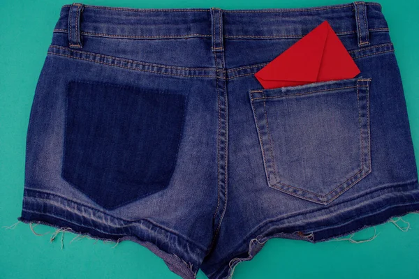 Blue denim shorts close up, back view on blue background, next to empty area. There is not enough pocket on jeans and red envelope in the second pocket.
