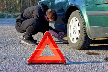 The guy in the black tracksuit put the jack under the car. In the foreground there is a warning sign.