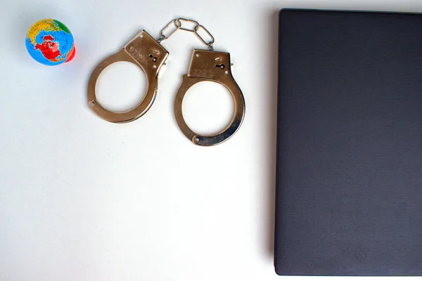 On a white table, the edge of a gray laptop, handcuffs and a globe. Near a copy space. Cybercrime concept.