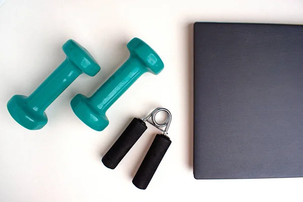 On a white table, the edge of a gray laptop, a wrist expander and two green dumbbells. Next to a copy space. The concept of workout online.