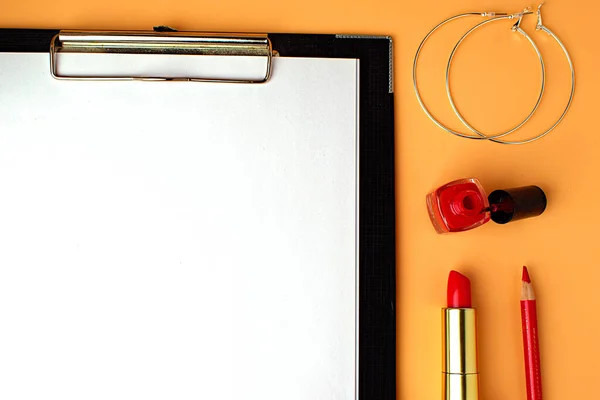 Writing pad with white sheet, next to red lipstick, red nail polish and round earrings. Notepad, to do list, get ready for date. A peach background. Copy space.