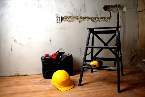 Black ladder, hard hat, ear protectors, protective gloves and tool case against the background of a concrete wall. Renovation concept.