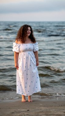 Kıvırcık saçlı bir kadın kumsalda huzurlu bir anın tadını çıkarır. Dalgaların gün batımında kıyıya vurması gibi çiçekli, dalgalı beyaz bir elbise giyer..