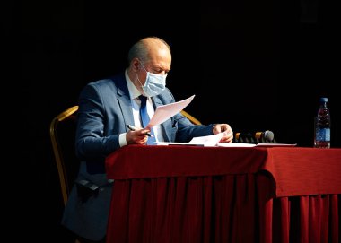 Şık ceketli ve koruyucu maskeli yaşlı bir adam kırmızı bir masa örtüsüyle masaya oturur ve kağıdı alır.
