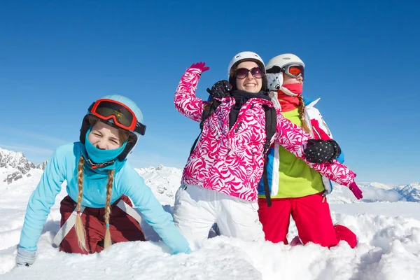 Three teenage girls on winter vacation in the mountains sitting in fresh snow look at the camera. Landscape with big blue sky and colorful clothes