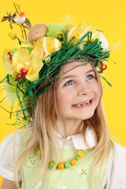 Mutlu kız Paskalya yumurtası avında saman yuvası saç stili, bahar çiçekleri, Paskalya yumurtaları ve tüylerden yapılmış. Çocuk gülümseyerek yan tarafa bakıyor. Sarı arkaplanda stüdyo portresi.