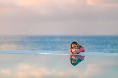 Kırmızı gömlekli genç bir kız, şafak vakti, deniz havuzu kenarında, sakin ve rahat bir şekilde, sudaki yansımaya bakıyor. Arka planda pembe bulutlar.