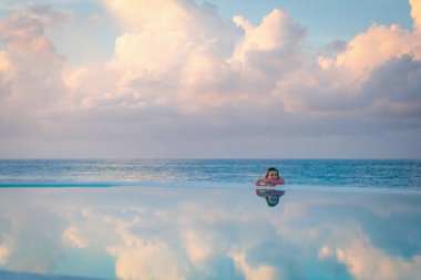 Kırmızı gömlekli genç bir kız, şafak vakti, deniz havuzu kenarında, sakin ve rahat bir şekilde, sudaki yansımaya bakıyor. Arka planda pembe bulutlar.