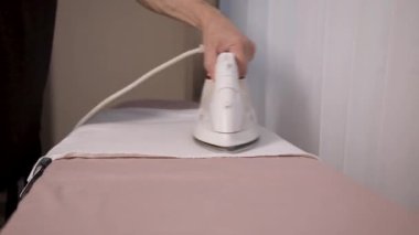 Ütü masasında beyaz elektrikli ütüyle çarşaf ütüleyen bir adam..